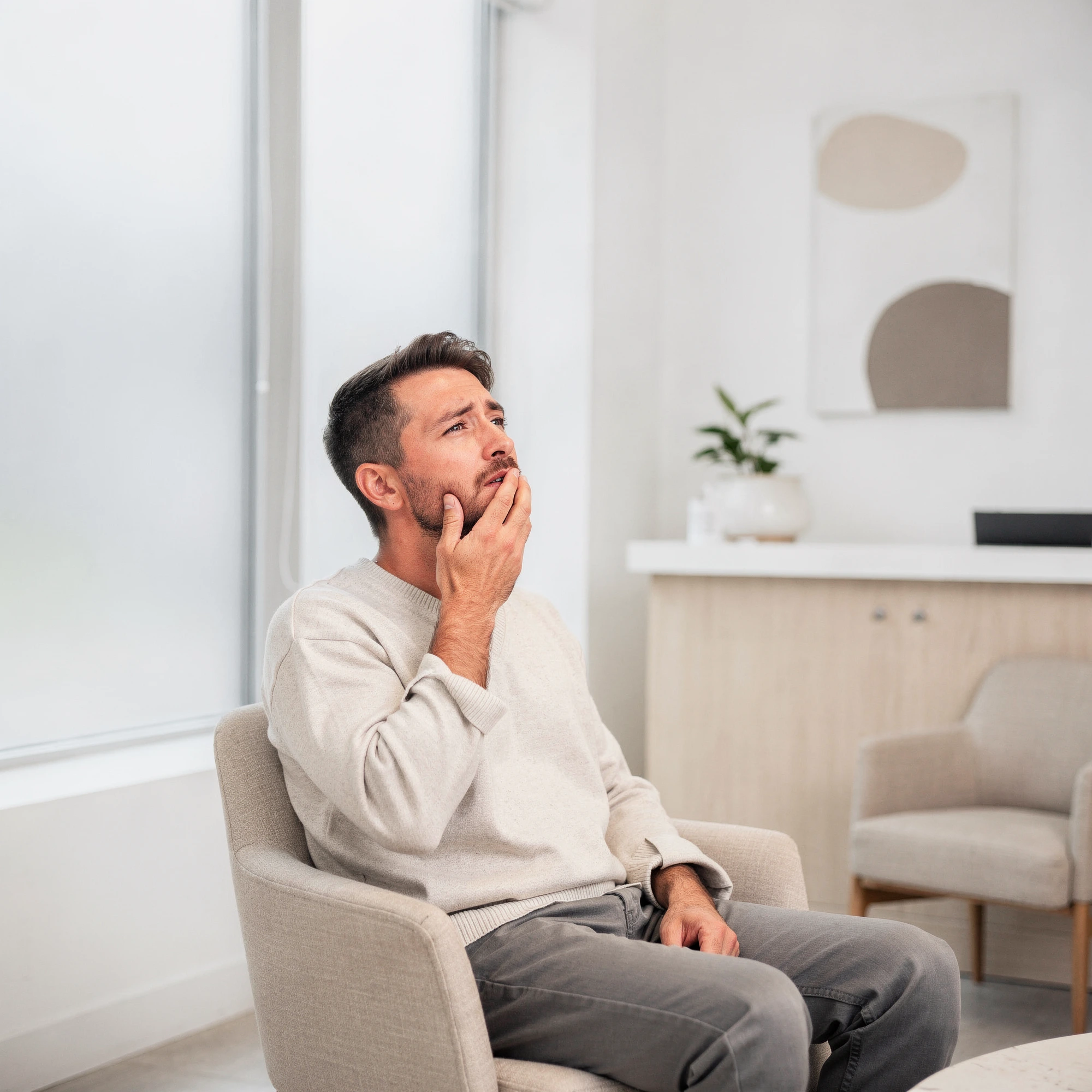 Man sitting in a waiting room holding his cheek in discomfort, indicating tooth pain